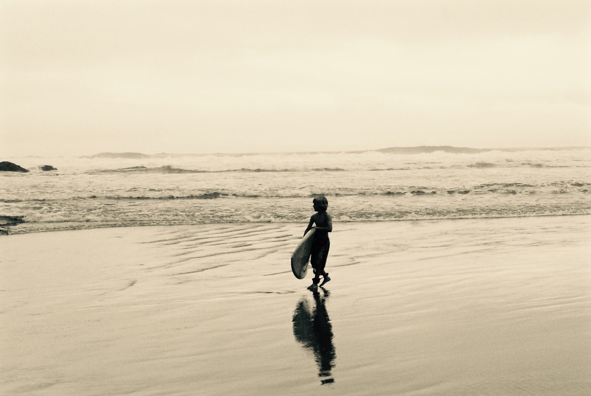 Child walking with a bodyboard on a calm beach, sepia-toned image.