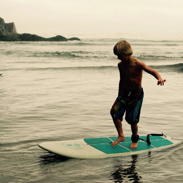 Child balancing on a surfboard in shallow waves near a rocky shore on a cloudy day.
