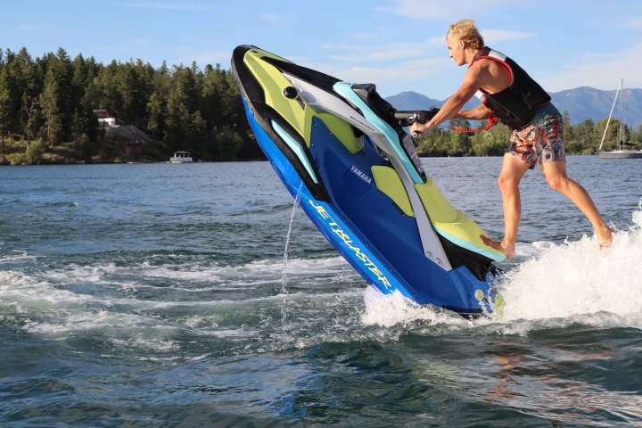 Person riding a jet ski on a lake, with trees and mountains in the background.