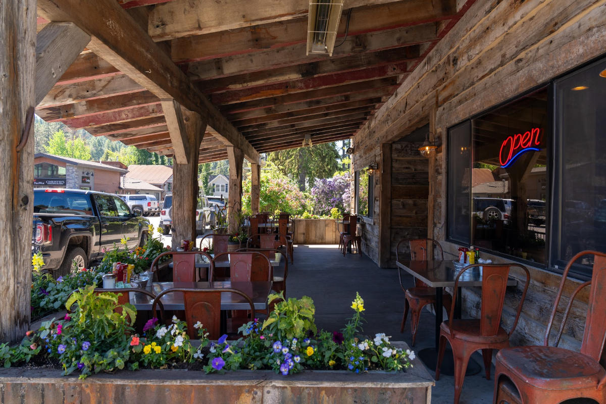 Wooden porch with tables, chairs, and potted flowers, under a roof with an 'Open' sign in the window.